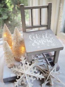 Top view of tan painted chair with "let it snow" and snow flakes painted in white on the seat of the chair. On the floor to the left of the chair are 3 off-white lit up trees and a white painted wooden snowflake.