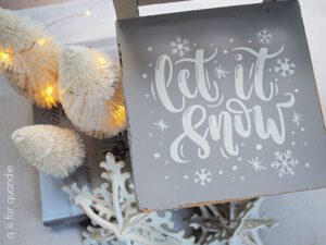 Top view of tan painted chair with "let it snow" and snow flakes painted in white on the seat of the chair. On the floor to the left of the chair are 3 off-white lit up trees and a white painted wooden snowflake.