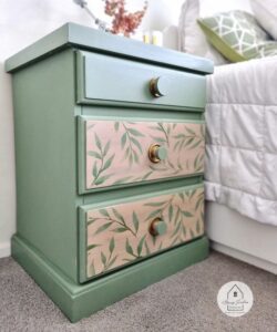 A green painted nightstand with brown painted drawers that have painted leaves on them sitting on a brown carpeted floor next to bed with a white bedspread and green throw pillow.