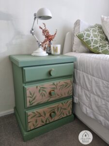 A green painted nightstand with brown painted drawers that have painted leaves on them sitting on a brown carpeted floor next to bed with a white bedspread and green throw pillow.