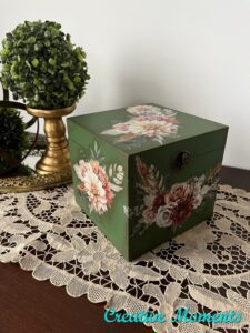 "After" photo of green painted wooden box with brown and white flowers on all sides sitting on top of a brown wooden table on a white lace doily.