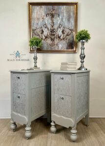 Gray painted nightstands with Royal Damask stencil sitting on a brown wooden floor in front of a white painted wall. Each nightstand has a silver plant holder with greenery coming out of it sitting on top. A brown painted picture is hanging on the wall.