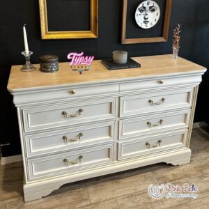 An off-white painted dresser with a painted wooden top sitting on a brown wooden floor, in front of a black painted wall. On top of the dresser are 4 different decorations, and two gold picture frames hang on the wall, the right one around a round clock that has a face on it.