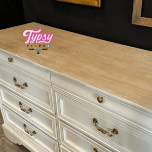 An off-white painted dresser with a painted wooden top sitting on a brown wooden floor, in front of a black painted wall.