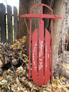 A red painted sled standing up on a ground covered with leaves, leaning up against a tree; the sled has stenciled words painted with white paint, The North Pole Bed & Breakfast Milk & Cookies Nightly