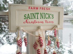 A white painted board that has red and white stockings hanging from it on white rope, the following words were stenciled with red and green paint: Farm Fresh Saint Nicks Christmas Trees