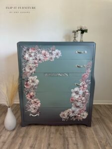 A gray painted chest of drawers with pink, white and gray florals along the edges sitting on a brown wooden floor in front of a white wall. On te floor to the left of the dresser is a white vase holding offwhite wheat grass