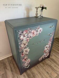 A gray painted chest of drawers with pink, white and gray florals along the edges sitting on a brown wooden floor in front of a white wall. On te floor to the left of the dresser is a white vase holding offwhite wheat grass