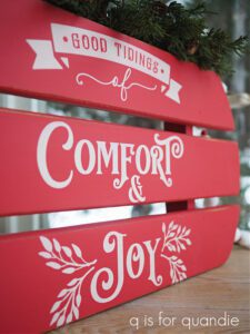 A red painted sled with white painted stenciled words Good Tidings, Comfort & Joy sitting on a brown wooden table