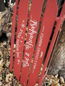 A red painted sled standing up on a ground covered with leaves, leaning up against a tree; the sled has stenciled words painted with white paint, The North Pole Bed & Breakfast Milk & Cookies Nightly