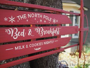 A red painted sled standing up on a ground covered with leaves, leaning up against a tree; the sled has stenciled words painted with white paint, The North Pole Bed & Breakfast Milk & Cookies Nightly