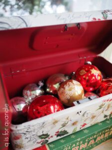 A red and white painted metal box with gold, silver and red christmas balls inside; the outside of the box has holly, berries and other christmas decorations on it.