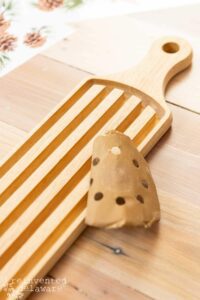 A rectangular wooden breadboard with a handle sitting on a wooden table. A round piece of sandpaper sits on top of the breadboard.