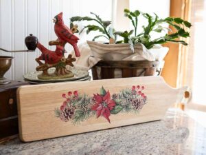 Rectangular wooden breadboard with a handle that has the Caroling Cardinals transfer applied to the center on a marble countertop with a statue of two cardinals and a green plant behind it