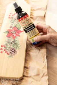 A hand holding a bottle of Easy Peasy Spray Wax over a rectangular wooden breadboard that has the Caroling Cardinals transfer applied to it. The breadboard is sitting on top of brown paper on a brown wooden table.