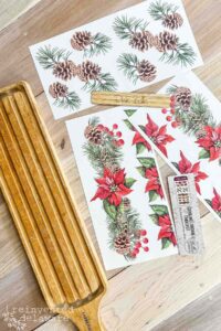 A wooden rectangular breadboard with a handle sits on a wooden tabletop with 3 sheets from the Caroling Cardinals transfer sitting on the table to the right of the breadboard.