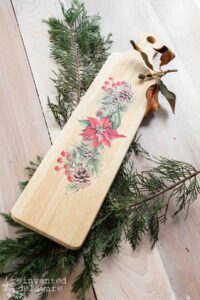 A rectangular wooden breadboard with a handle that has the Caroling Cardinals transfer applied sits on top of a pine branch on top of a wooden table.+