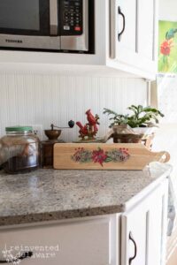 Rectangular wooden breadboard with a handle that has the Caroling Cardinals transfer applied to the center on a marble countertop with a statue of two cardinals and a green plant behind it